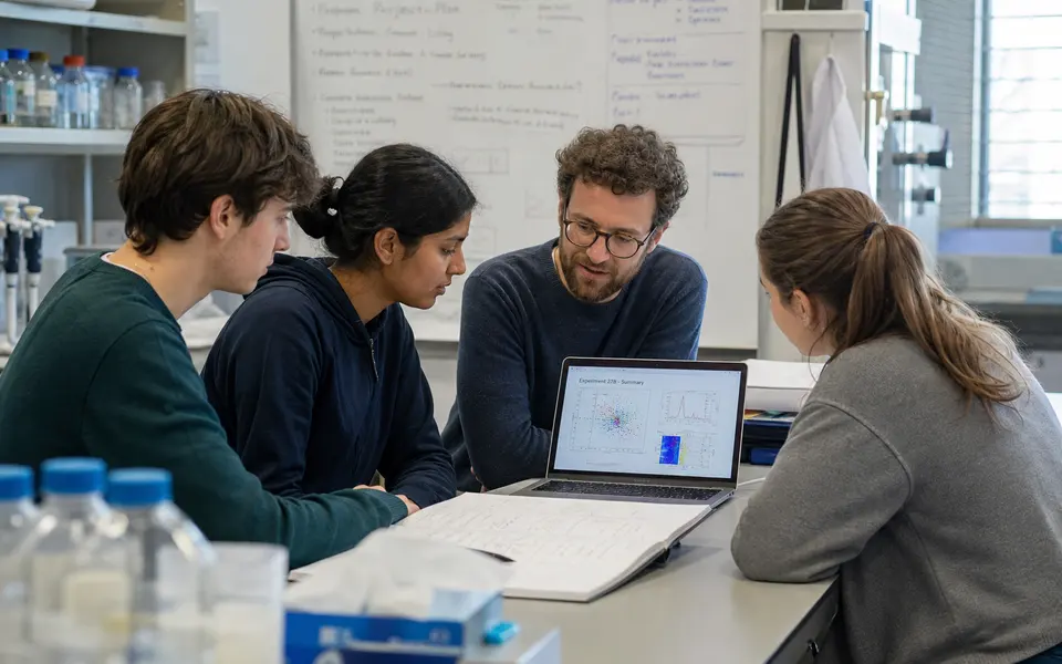 A small group of diverse undergraduate students, including an Indian student, discussing results with a supervisor around a lab bench or shared workspace.