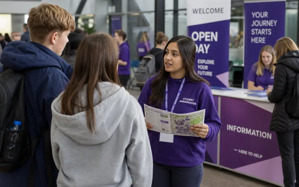 Indian student working as a student ambassador at a UK university open day, talking to visitors near an information desk.